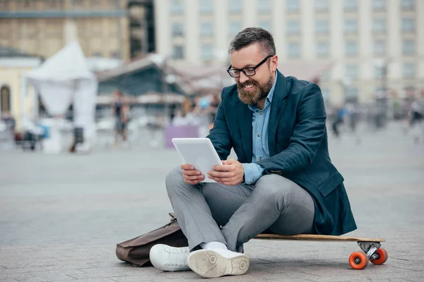 Sorrindo barbudo homem usando laptop e sentado em longboard na cidade — Fotografia de Stock