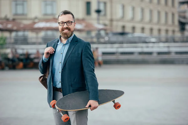 Stylish bearded man with leather bag and skateboard in city — Stock Photo