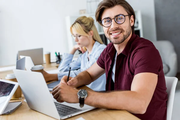 Colegas que trabajan en el proyecto de inicio en la oficina y el hombre sonriente mirando a la cámara - foto de stock