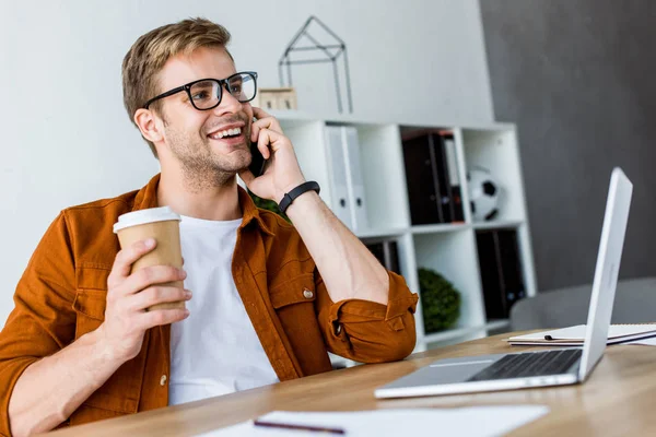 Lächelnder gutaussehender Geschäftsmann, der im Büro an einem Startup-Projekt arbeitet und per Smartphone spricht — Stockfoto