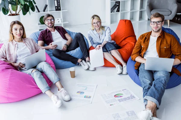 Colegas multiculturales que trabajan en el proyecto de puesta en marcha en la oficina y sentarse en sillas de la bolsa de frijol con gadgets — Stock Photo