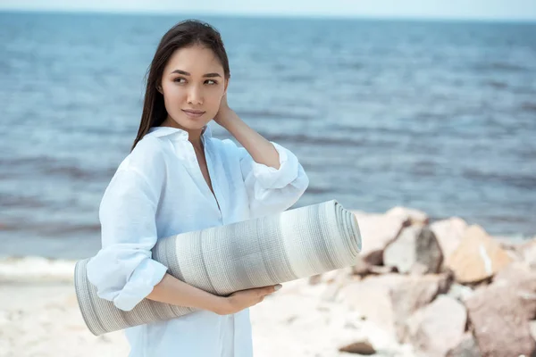 Attrayant asiatique femme tenant tapis de yoga et regardant loin par la mer — Photo de stock