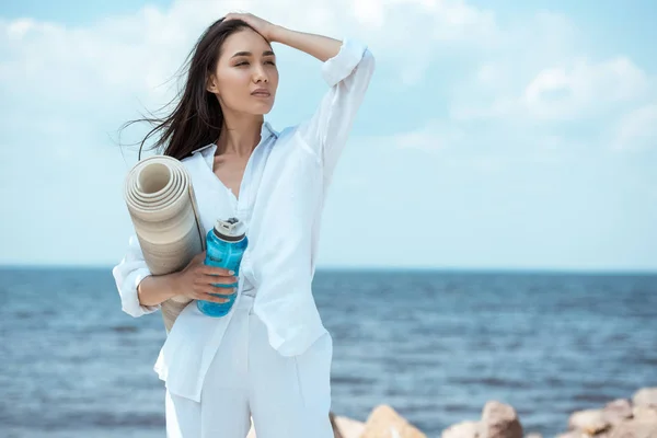 Asiatische junge Frau hält Sportflasche mit Wasser und Yogamatte am Meer — Stockfoto