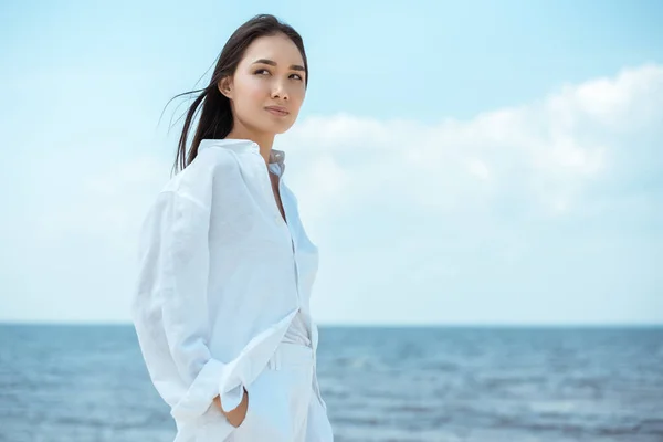 Junge asiatische Frau mit Händen in Taschen, die vom Meer wegschauen — Stockfoto