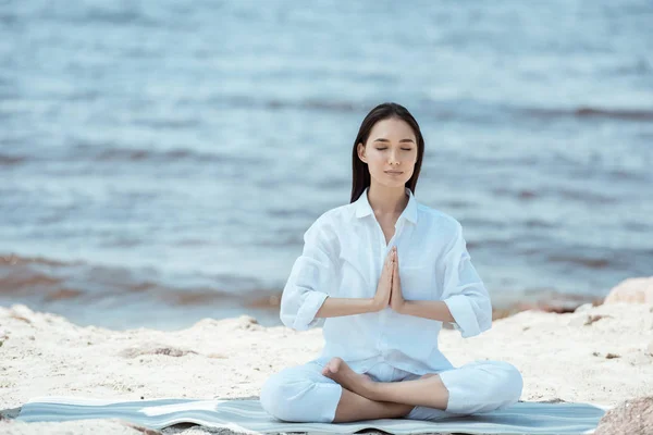 Attractive asian woman meditating in anjali mudra (salutation seal) pose on yoga mat by sea — Stock Photo