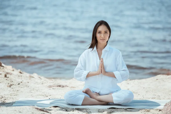 Joven asiático mujer en anjali mudra (salutation seal) pose en yoga mat por mar - foto de stock
