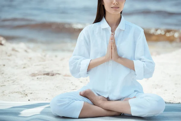Cropped image of woman meditating in anjali mudra (salutation seal) pose on yoga mat by sea — Stock Photo