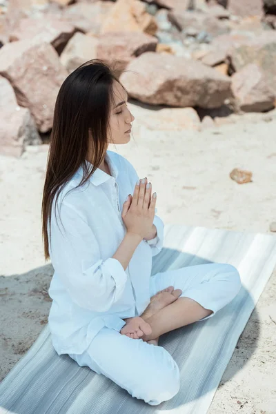 Vista de ángulo alto de la mujer asiática enfocada meditando con los ojos cerrados en la pose de anjali mudra en la esterilla de yoga - foto de stock