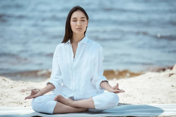 Focused woman meditating in ardha padmasana (half lotus pose) on yoga mat by sea — Stock Photo