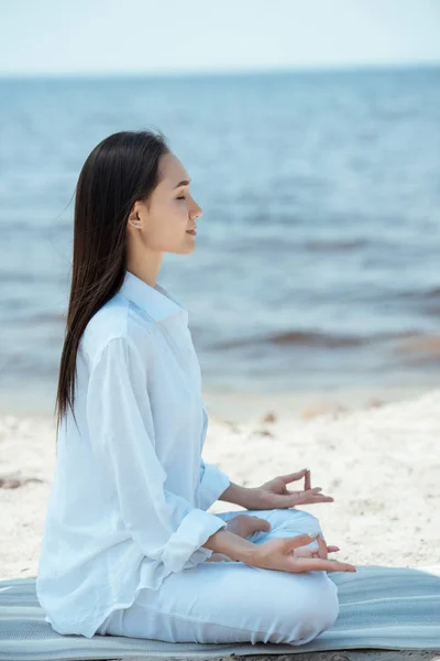 Side view of young asian woman in ardha padmasana (half lotus pose) on yoga mat by sea — Stock Photo