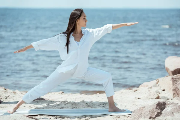Mujer asiática joven concentrada practicando yoga en virabhadrasana (pose guerrera II) sobre una esterilla frente al mar - foto de stock