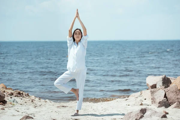 Азиатская женщина, стоящая в asana vrikshasana (поза дерева) на пляже на море — стоковое фото