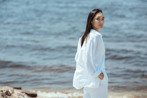 Приваблива азіатська жінка з руками в кишенях дивиться на камеру біля моря — стокове фото