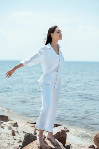 Feliz mulher asiática com olhos fechados desfrutando do mar e de pé com os braços estendidos na praia — Fotografia de Stock