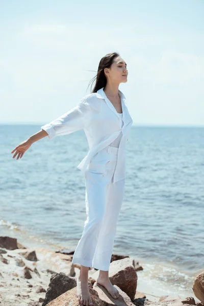 Mujer asiática feliz con los ojos cerrados disfrutando del mar y de pie con los brazos extendidos en la playa - foto de stock