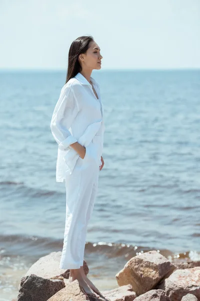 Side view of attractive asian woman with hand in pocket standing on beach by sea — Stock Photo