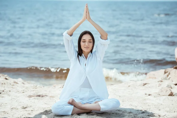 Focused asian young woman meditating in lotus position and doing namaste mudra gesture above head by sea — Stock Photo
