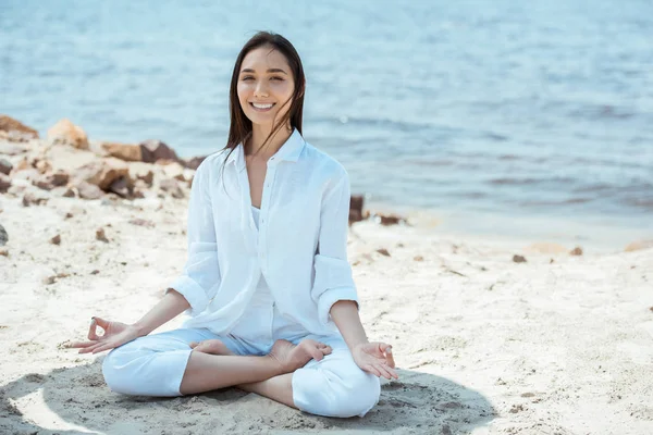 Sonriente asiático mujer en ardha padmasana (medio loto pose) en playa por mar - foto de stock