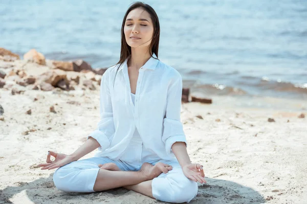 Mujer asiática con los ojos cerrados meditando con akash mudra gesto en posición de loto en la playa - foto de stock