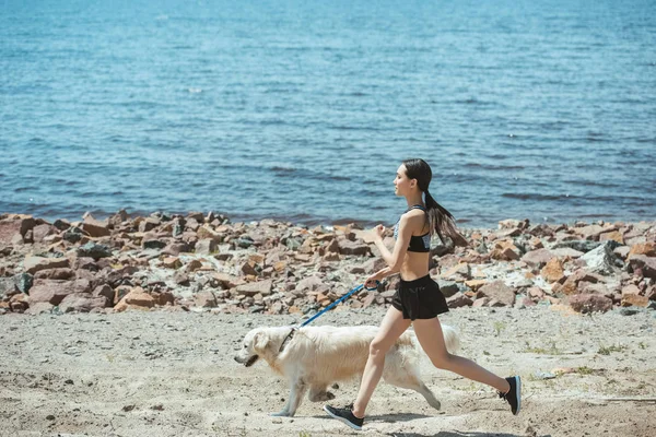 Side view of asian sportswoman jogging with dog on beach — Stock Photo