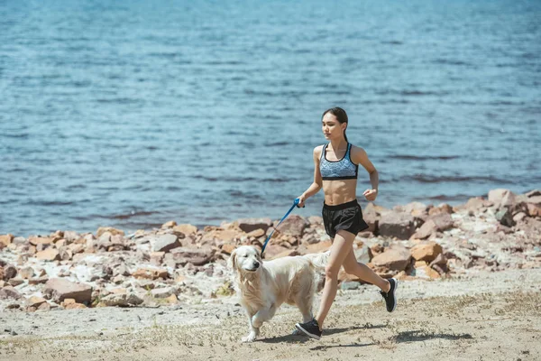 Young asian sportswoman running with golden retriever on beach — Stock Photo