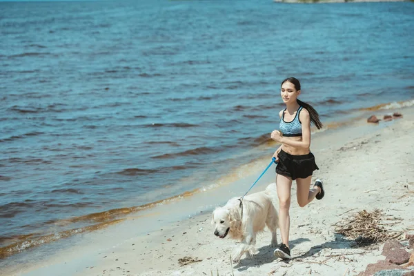 Young asian sportswoman running with golden retriever near sea — Stock Photo