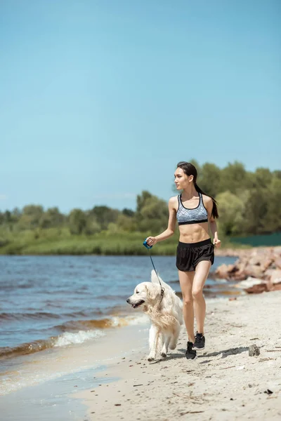 Attractive young asian sportswoman running with golden retriever near sea — Stock Photo