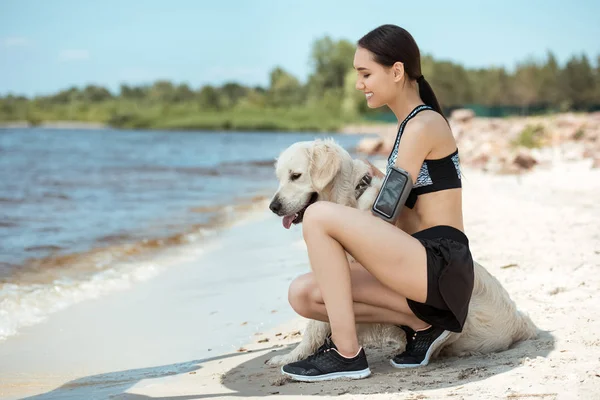 Feliz asiático esportista com smartphone em execução braçadeira caso abraçando golden retriever na praia — Fotografia de Stock