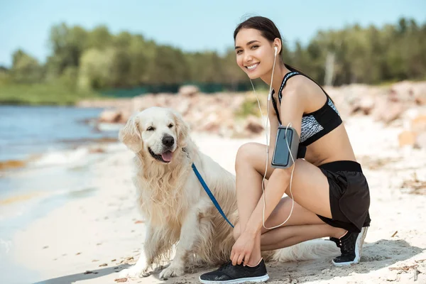 Smiling asian sportswoman in earphones with smartphone in running armband case tying shoelaces near golden retriever on beach — Stock Photo