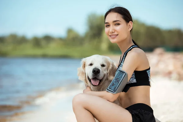Smiling asian sportswoman with smartphone in running armband case embracing golden retriever — Stock Photo