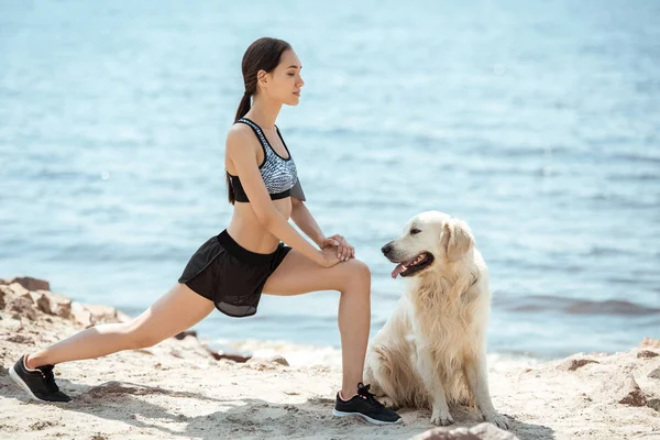 Side view of asian female athlete doing lunges near golden retriever on beach — Stock Photo