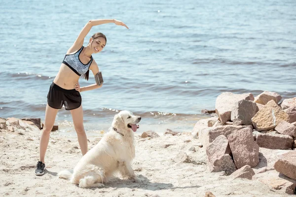 Smiling asian sportswoman with smartphone in running armband case doing exercise while her golden retriever sitting near on beach — Stock Photo