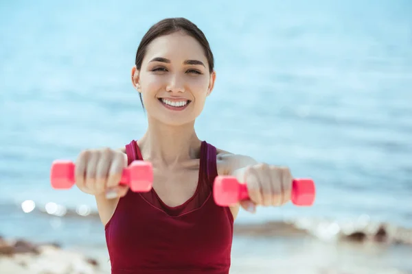 Sorridente asiatica sportiva che si esercita con i manubri di fronte al mare — Foto stock