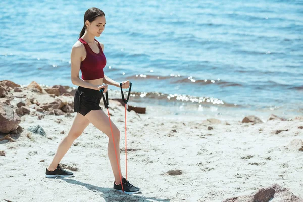 Vista laterale della giovane sportiva che fa esercizio con fascia elastica sulla spiaggia — Foto stock
