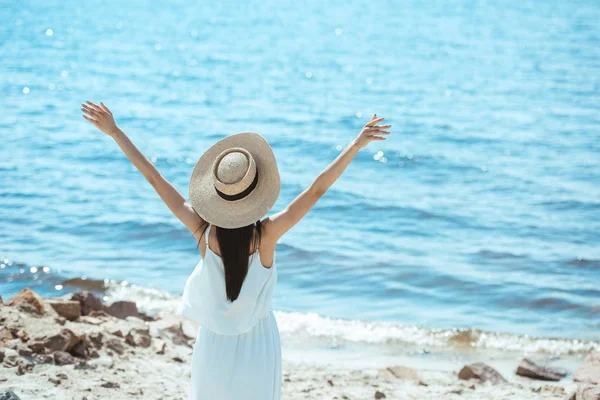 Vue arrière de la femme en chapeau de paille et robe blanche debout avec de larges bras par la mer — Photo de stock