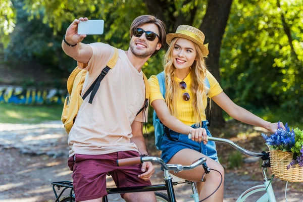 Sonriente pareja enamorada de las bicicletas tomando selfie en el parque en el día de verano - foto de stock