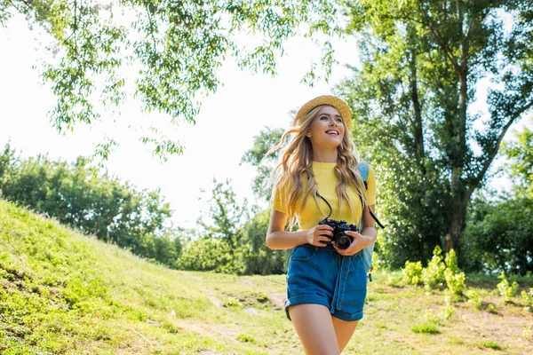 Giovane donna sorridente in cappello con macchina fotografica passeggiando nel parco estivo — Foto stock