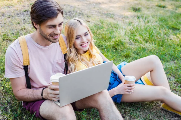 Jeune couple souriant avec café pour aller utiliser un ordinateur portable dans le parc — Photo de stock
