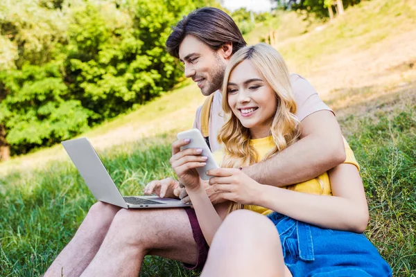 Jovem casal com laptop e smartphone descansando na grama verde no parque — Fotografia de Stock