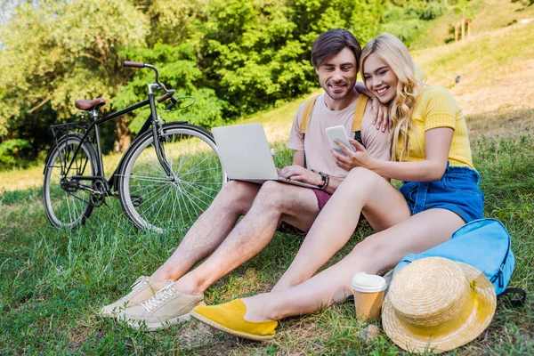 Pareja joven con ordenador portátil y smartphone descansando sobre hierba verde en el parque - foto de stock