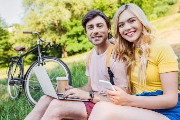 Coppia sorridente con computer portatile e smartphone poggiato su erba verde nel parco — Foto stock