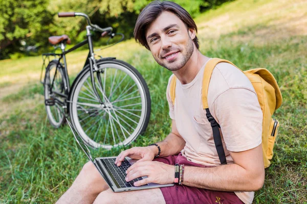 Vista lateral del hombre sonriente con la mochila, el ordenador portátil y la bicicleta cerca sentado en la hierba verde en el parque - foto de stock