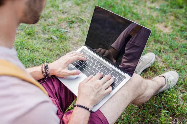 Vista parcial del hombre usando el ordenador portátil mientras está sentado en la hierba verde en el parque - foto de stock