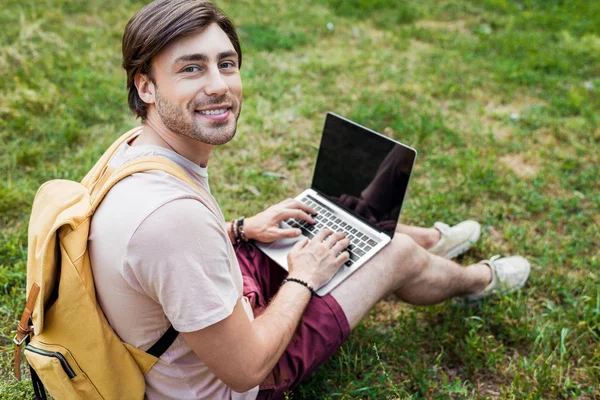 Vista laterale di uomo sorridente con zaino e computer portatile seduto su erba verde nel parco — Foto stock