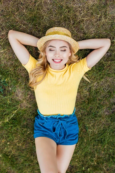 Overhead view of smiling young woman in hat resting on green grass in park — Stock Photo