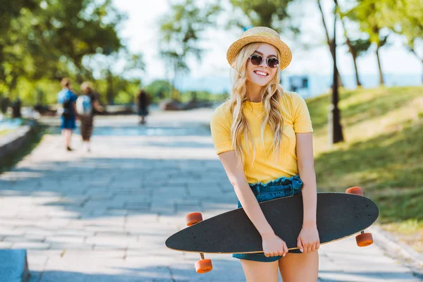 Portrait of smiling blond woman in sunglasses and hat with longboard in hands on street — Stock Photo