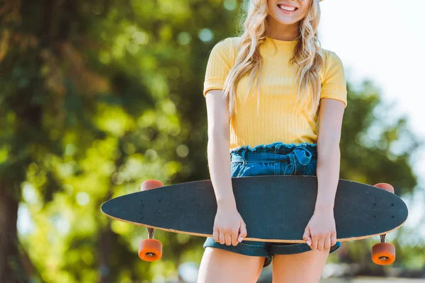 Vista parcial de mulher loira sorridente com longboard de pé na rua — Fotografia de Stock