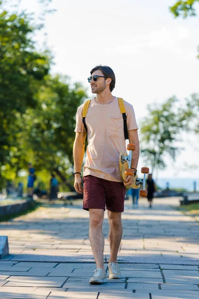 Junger Mann mit Sonnenbrille, Rucksack und Longboard läuft auf der Straße — Stockfoto