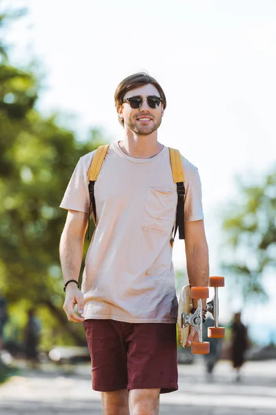 Ritratto di giovane in occhiali da sole con zaino e longboard che cammina per strada — Foto stock