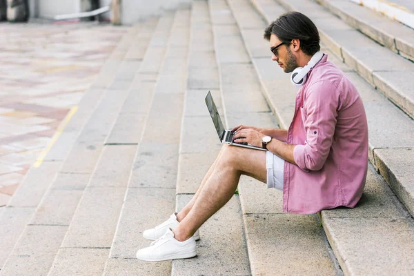 Vue latérale de l'homme en lunettes de soleil avec casque sur le cou à l'aide d'un ordinateur portable tout en étant assis sur les marches sur la rue — Photo de stock